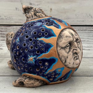 May include: A ceramic celestial sculpture featuring a blue sphere with cut-out holes, adorned with gold and turquoise accents. A carved face of the moon is on the front. The sculpture has a textured, earthy base and top.