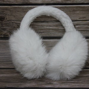 May include: White faux fur earmuffs with a knitted headband. The ear coverings are fluffy and round, designed to keep ears warm. The background is a wooden surface.