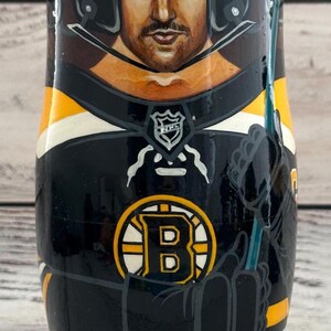May include: A hand-painted wooden nesting doll featuring a hockey player in a black and gold Boston Bruins uniform. The doll has a portrait of the player's face, a black helmet with the CCM logo, and the words "BOSTON BRUINS" at the base.