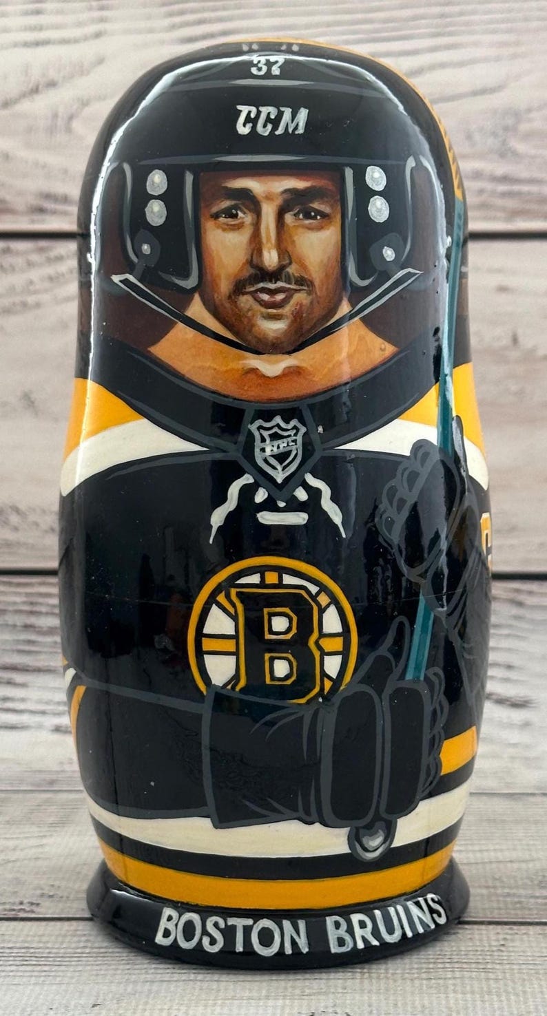 May include: A hand-painted wooden nesting doll featuring a hockey player in a black and gold Boston Bruins uniform. The doll has a portrait of the player's face, a black helmet with the CCM logo, and the words "BOSTON BRUINS" at the base.