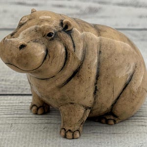 May include: A detailed, tan-colored hippo figurine. The sculpture has a rounded body, small ears, and a smiling expression. The hippo is resting on all four legs. The figurine is approximately 5 inches long.