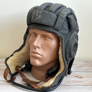 Può includere: Un casco da carrista imbottito grigio scuro con esterno trapuntato, una toppa rettangolare frontale con il numero 15 e una fodera in pelliccia sintetica color crema. Il casco include sottogola in pelle marrone e un bordo in gomma nera.