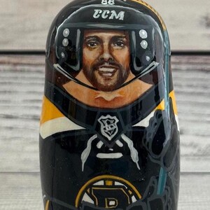 May include: A hand-painted wooden nesting doll featuring a hockey player in a black and gold uniform. The doll has a portrait of the player's face, a helmet with the text "CCM" and the number "86". The doll is a collectible item.