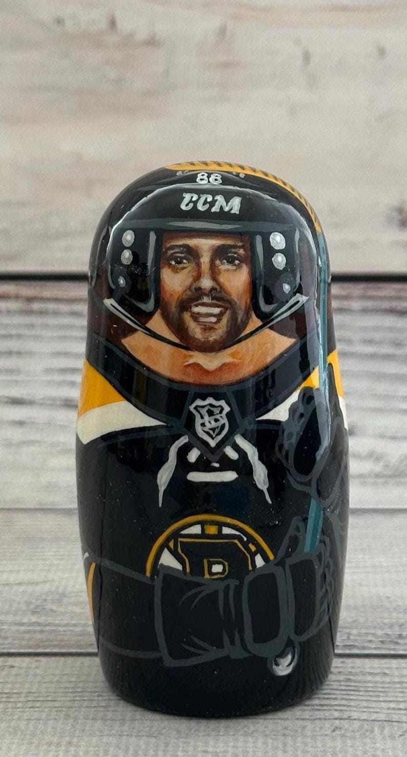 May include: A hand-painted wooden nesting doll featuring a hockey player in a black and gold uniform. The doll has a portrait of the player's face, a helmet with the text "CCM" and the number "86". The doll is a collectible item.