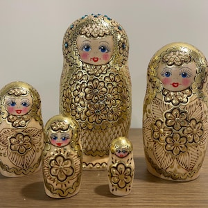 May include: A set of five gold-painted nesting dolls, each with a floral design and a painted face with blue eyes and red lips. The dolls are arranged from largest to smallest, showcasing the traditional Russian Matryoshka style.