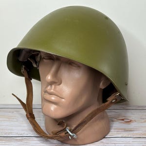 Puede incluir: Casco militar verde oliva con barboquejo de cuero. El casco se exhibe sobre una cabeza de maniquí. El casco tiene forma redondeada y acabado mate. El barboquejo es marrón y se asegura con herrajes metálicos.