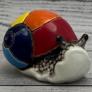 Puede incluir: Figura de caracol de cerámica con una concha colorida. La concha está segmentada en secciones amarillas, naranjas, rojas y azules. El cuerpo del caracol es blanco con un patrón texturizado y tiene antenas negras.