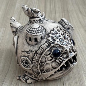 Puede incluir: Escultura de cerámica detallada de una criatura con forma de pez con elementos arquitectónicos. La escultura es principalmente blanquecina con detalles en azul oscuro para los ojos. Presenta detalles intrincados.