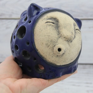 Könnte beinhalten: Eine skurrile Keramikskulptur in Form eines Gesichts, mit marineblauem Äußeren und cremefarbenem Gesicht. Die Skulptur hat eine Reihe kreisförmiger Aussparungen und kleine ohrartige Vorsprünge. Das Gesicht hat geschlossene Augen und einen kleinen, offenen Mund.