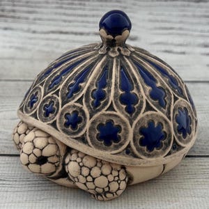 May include: A ceramic turtle figurine with a detailed, ornate shell. The shell is a deep blue with a decorative pattern, and the turtle's body is a cream color. The figurine has a lid with a small blue knob.