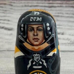 May include: A hand-painted wooden nesting doll featuring a hockey player in a black and gold uniform. The doll has a painted face and a helmet with the text "CCM" and the number "73". The doll is a collectible item.