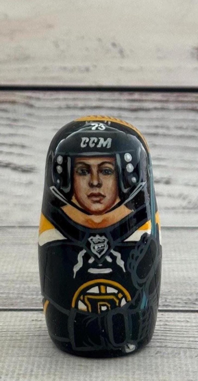 May include: A hand-painted wooden nesting doll featuring a hockey player in a black and gold uniform. The doll has a painted face and a helmet with the text "CCM" and the number "73". The doll is a collectible item.