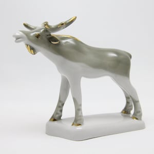 Puede incluir: Una figura de cerámica brillante de un alce de pie sobre una base rectangular. El alce está coloreado en tonos gris claro y blanco, con detalles en oro metálico en la cornamenta, el cuello y los cascos, con un acabado suave y brillante.