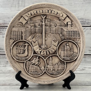 May include: A beige decorative plate featuring a detailed relief of Kyiv, Ukraine. The plate includes cityscapes, monuments, and the Ukrainian word for Ukraine. It is displayed on a black stand.