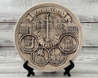 May include: A beige decorative plate featuring a detailed relief of Kyiv, Ukraine. The plate includes cityscapes, monuments, and the Ukrainian word for Ukraine. It is displayed on a black stand.