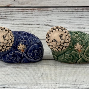 Op de afbeelding: Twee keramische leeuwenbeeldjes. Eén is donkerblauw, de ander groen. Beide hebben een leeuwenkop met een gedetailleerde manen en een bloemmotief op hun lichaam. Op een licht houten oppervlak.