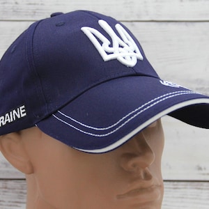 Puede incluir: Gorra de béisbol azul marino con un emblema blanco bordado y la palabra "UKRAINE" en el lateral. La gorra tiene una visera curva con ribete y costuras blancas. La gorra se muestra en una cabeza de maniquí.