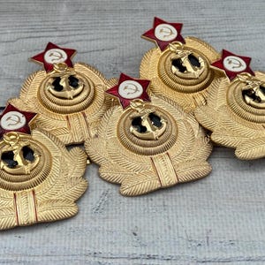 Può includere: Cinque distintivi in metallo color oro con stelle rosse e bianche e ancore. I distintivi hanno un design a corona di alloro.