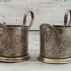 May include: Two antique silver-colored cup holders with handles. Each holder features an embossed design with floral and geometric patterns. The cup holders are cylindrical with a circular base and a handle attached to the side.