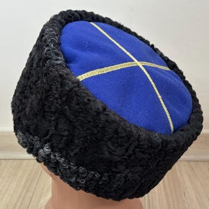 Könnte beinhalten: Ein traditioneller Kosakenhut mit blauem Oberteil und schwarzem Pelzbesatz. Das blaue Oberteil hat ein goldenes Kreuzmuster. Der Hut wird auf einem Schaufensterkopf präsentiert.