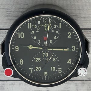 Op de afbeelding: Een zwarte ronde cockpitklok voor de luchtvaart met een donkere wijzerplaat, witte cijfers en twee sub-wijzerplaten. De bovenste sub-wijzerplaat bevat Cyrillische tekst en een rood vierkant, terwijl de onderste de Cyrillische tekst voor seconden toont. Inclusief een rode knop en een zilveren knop.