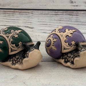 May include: Two ceramic snail figurines with intricate gold designs on their shells. One snail has a green shell and the other has a purple shell.