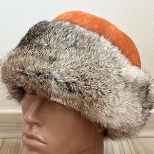 Könnte beinhalten: Ein Winterhut mit Pelzbesatz und einer Krone aus gebranntem orangefarbenem Wildleder. Das Fell des Hutes ist eine Mischung aus Grau und Braun, wodurch ein strukturiertes Aussehen entsteht. Der Hut ist zum Schutz vor Kälte konzipiert.