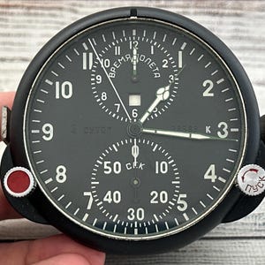 Op de afbeelding: Een zwarte ronde cockpitklok met een matte afwerking, voorzien van witte Arabische cijfers en indexen. De wijzerplaat bevat twee sub-wijzerplaten, Cyrillische tekst en twee zijknoppen, één met een rode bovenkant en één met witte tekst.