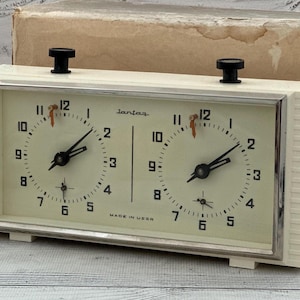 May include: A vintage cream-colored analog chess clock with two circular clock faces, black numerals, and black hands. The device features two black top-mounted buttons, a silver-toned metal frame, and the text Jantar and Made in USSR on the face.