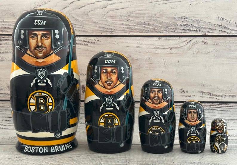 May include: A set of five nesting dolls, each painted to resemble a hockey player in a black, yellow, and white uniform. The largest doll reads "BOSTON BRUINS" at the base. Each doll has a hockey helmet and the number 37, 69, 66, 72, and 75.