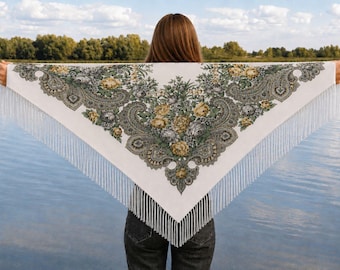 May include: A white triangular shawl with a floral and paisley design in shades of green, yellow, and gray. The shawl has a fringe along the edges. The shawl is held up against a backdrop of water and trees.
