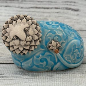 Puede incluir: Figura de león de cerámica en posición reclinada. La cabeza del león es blanquecina con detalles de pelaje, y el cuerpo es azul claro con un patrón arremolinado. Una pequeña flor de lis está en el lateral. La figura está sobre una superficie de madera clara.