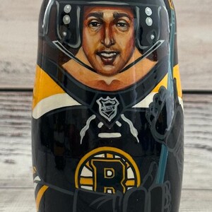 May include: A hand-painted wooden nesting doll featuring a hockey player in a black and gold uniform. The doll has the number 63 and the CCM logo on the helmet. The Boston Bruins logo is visible on the front.