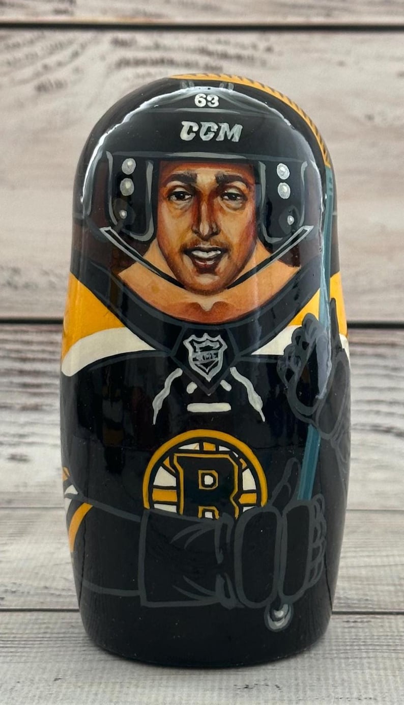 May include: A hand-painted wooden nesting doll featuring a hockey player in a black and gold uniform. The doll has the number 63 and the CCM logo on the helmet. The Boston Bruins logo is visible on the front.