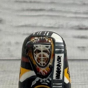 May include: A hand-painted, oval-shaped stone featuring a hockey goalie in a black, yellow, and white uniform. The goalie's mask is visible, along with the word "WARRIOR" on the side. The design includes the letter "P" and the team's logo.