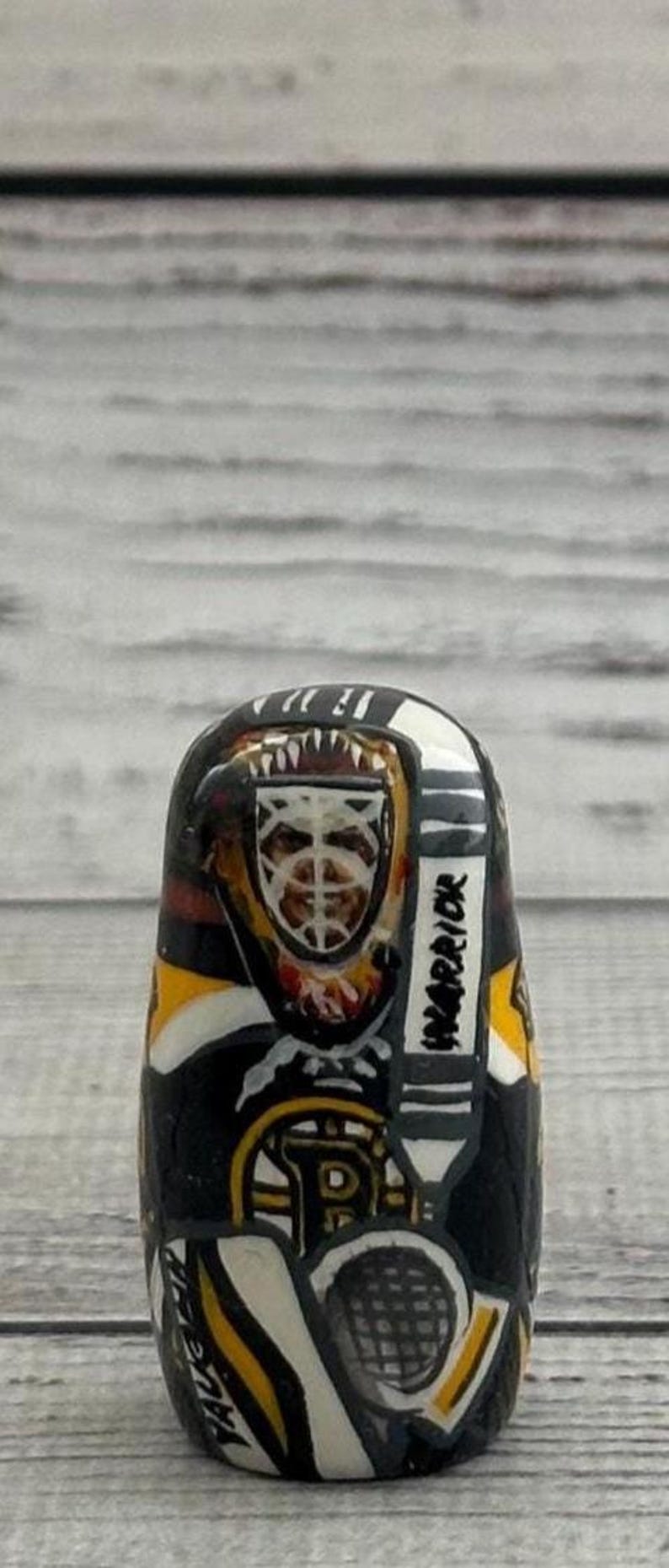 May include: A hand-painted, oval-shaped stone featuring a hockey goalie in a black, yellow, and white uniform. The goalie's mask is visible, along with the word "WARRIOR" on the side. The design includes the letter "P" and the team's logo.