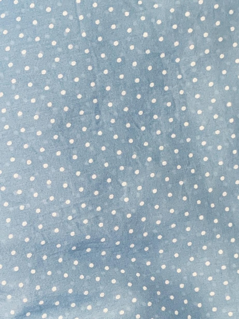 May include: A light blue fabric featuring a repeating pattern of small, solid white polka dots. The material has a subtle, slightly mottled texture with variations in the blue tone, creating a soft, consistent appearance across the surface.