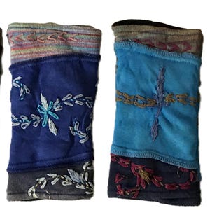 Op de afbeelding: Vier kleurrijke armwarmers met geborduurde bloemen- en geometrische patronen. De armwarmers hebben verschillende kleuren, waaronder blauw, turquoise en grijs, met decoratieve stiksels en een geribbelde manchet.