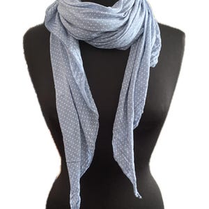 May include: A light blue scarf featuring a repeating pattern of small white polka dots. The scarf is draped in multiple layers around the neck, with two long ends hanging down the front, showcasing the soft, lightweight fabric texture.