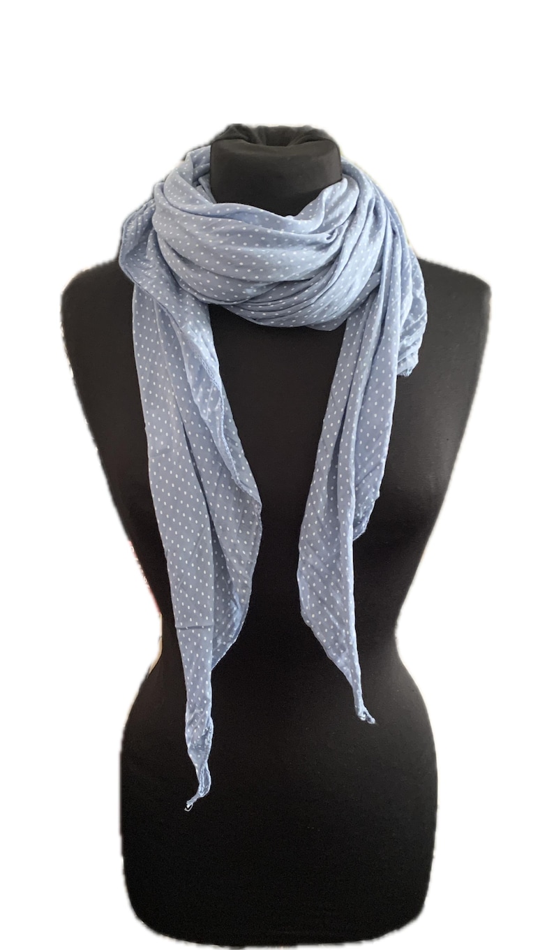 May include: A light blue scarf featuring a repeating pattern of small white polka dots. The scarf is draped in multiple layers around the neck, with two long ends hanging down the front, showcasing the soft, lightweight fabric texture.