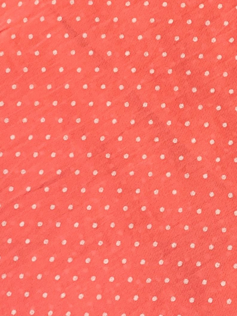 May include: A coral-colored fabric featuring a repeating pattern of small, white, circular polka dots. The material has a visible, slightly textured weave, creating a consistent and uniform appearance across the entire surface of the textile.
