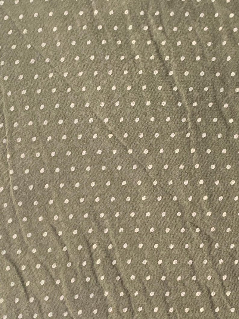 May include: A sage green fabric featuring a repeating pattern of small, white, circular polka dots. The material has a slightly textured, woven appearance with visible folds and creases running diagonally across the surface of the textile.