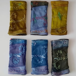 Puede incluir: Seis puños de tela con diferentes diseños de bordado. Los puños están hechos de un patchwork de telas azules, verdes y marrones. El bordado está en tonos de verde y azul.