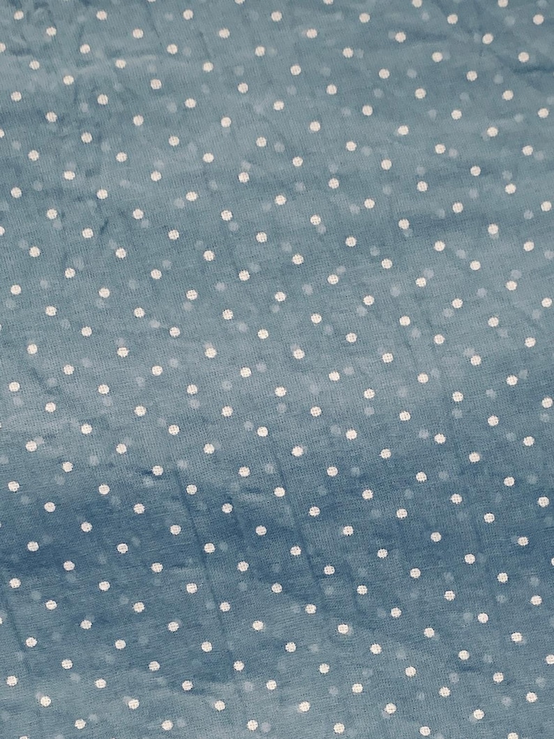 May include: A blue fabric featuring a repeating pattern of small, white, circular polka dots distributed evenly across the surface. The material has a slightly textured appearance with a consistent weave, showcasing a classic and simple design.