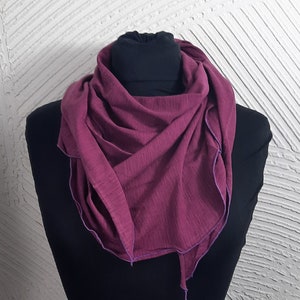 Puede incluir: Una bufanda de color burdeos oscuro con ribete morado. La bufanda está hecha de un tejido suave y de punto y está doblada y expuesta en un maniquí negro.