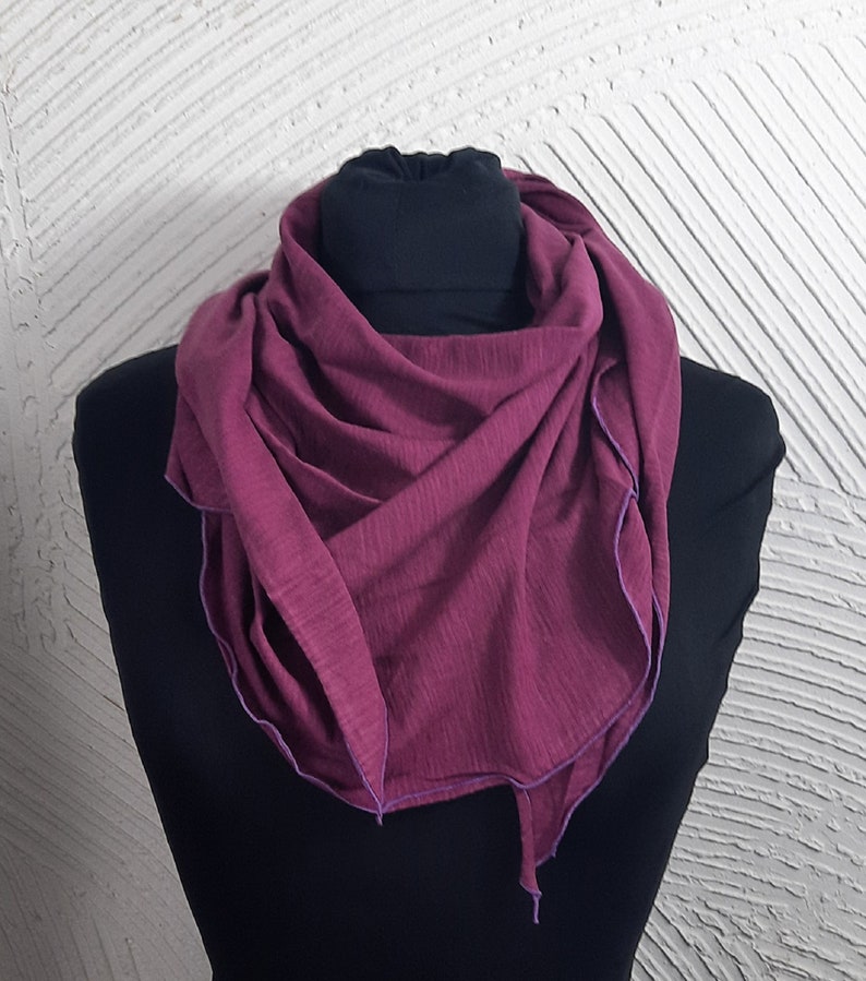 K&ouml;nnte beinhalten: Ein tiefroter Schal mit lilafarbener Einfassung. Der Schal ist aus weichem, gestricktem Stoff gefertigt und auf einer schwarzen Puppe gefaltet und ausgestellt.