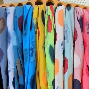 Può includere: Una collezione di sciarpe colorate appese a un supporto in legno. Le sciarpe presentano motivi astratti con cerchi e macchie in tonalità di blu, giallo, rosa e verde. Il tessuto appare leggero e fluido.