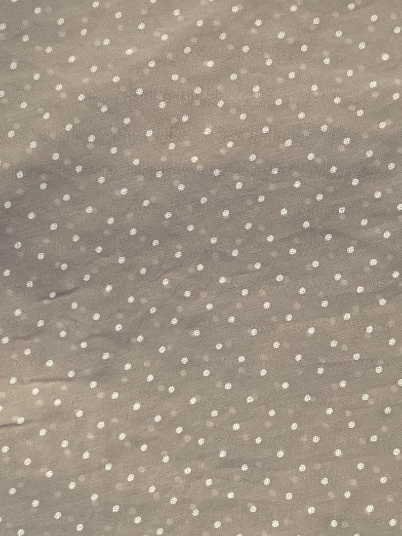 May include: A sheer, light taupe fabric featuring a repeating pattern of small, evenly spaced white polka dots. The material has a delicate, semi-transparent quality with a subtle texture visible across the entire surface of the textile.