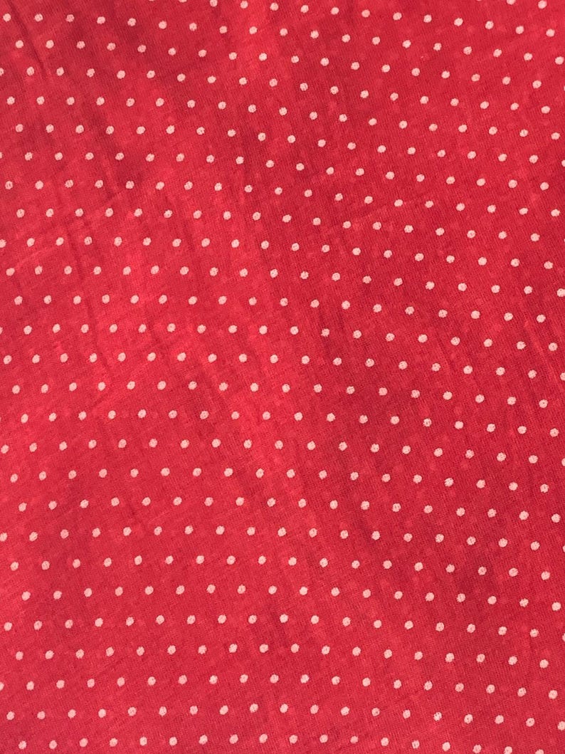 May include: A vibrant red fabric featuring a repeating pattern of small, evenly spaced white polka dots. The material has a slightly textured surface with a consistent weave, creating a uniform and classic dotted design across the entire visible area.