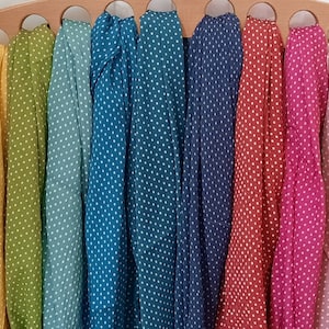 May include: A collection of eight lightweight scarves in yellow, olive green, teal, blue, navy, red, magenta, and light pink, each featuring a white polka dot pattern. The scarves are draped over a wooden hanger with eight circular cutouts.